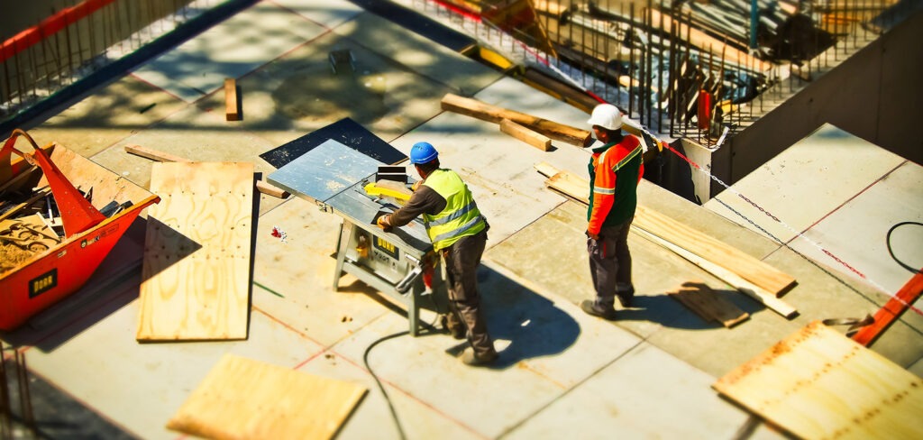 two construction workers at a construction site cutting sheets of wood with a power saw