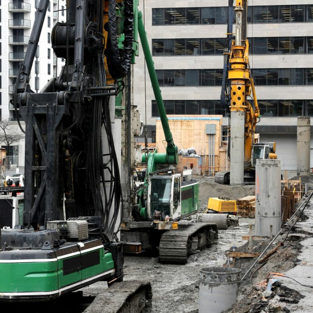 Three augers on an active construction site drilling into the ground to create the foundation for a new building.