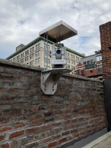 An automated motorized total station mounted on a bracket attached to a brick wall monitors a structure located in a construction zone for movement.