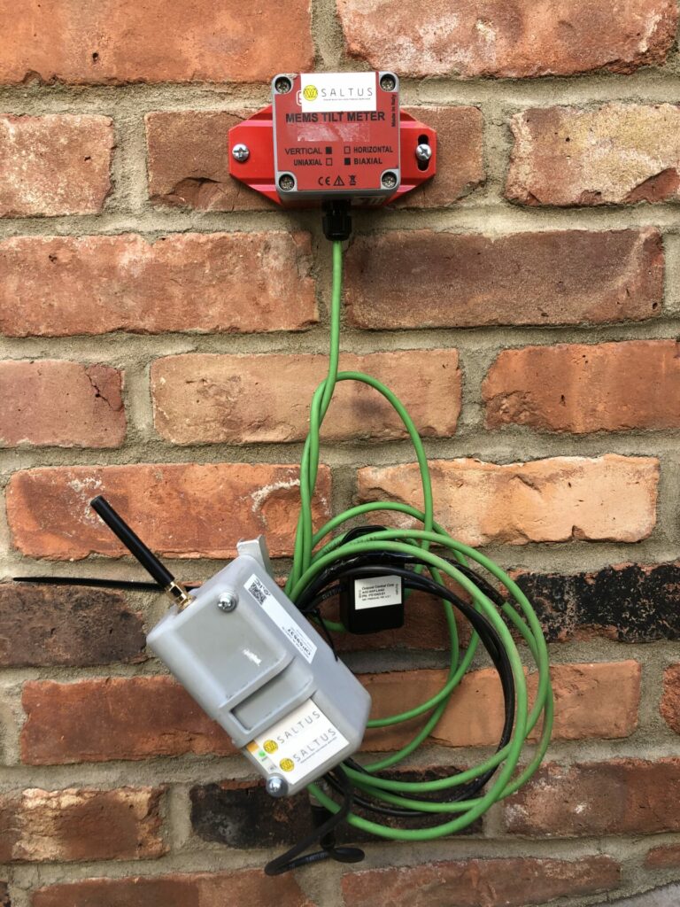 A tiltmeter installed on the brick wall of a building located near an active construction site.