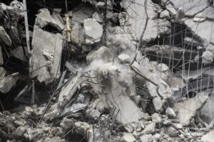 Demolition site with crumbling concrete slabs, exposed rebar, and airborne dust rising from collapsed building debris.